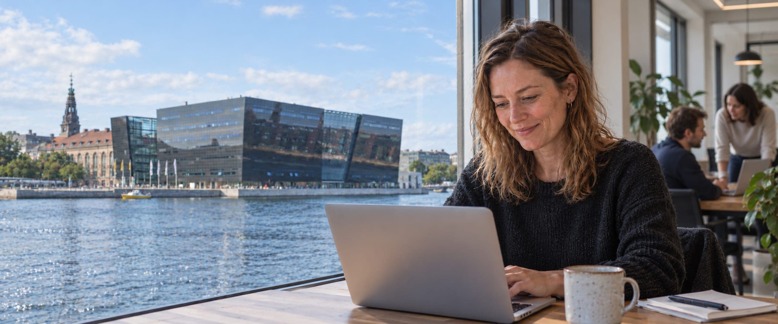 En smilende kvinde arbejder ved sin laptop i et kontor med udsigt over havnen til Den Sorte Diamant i København. To kolleger taler sammen i baggrunden.